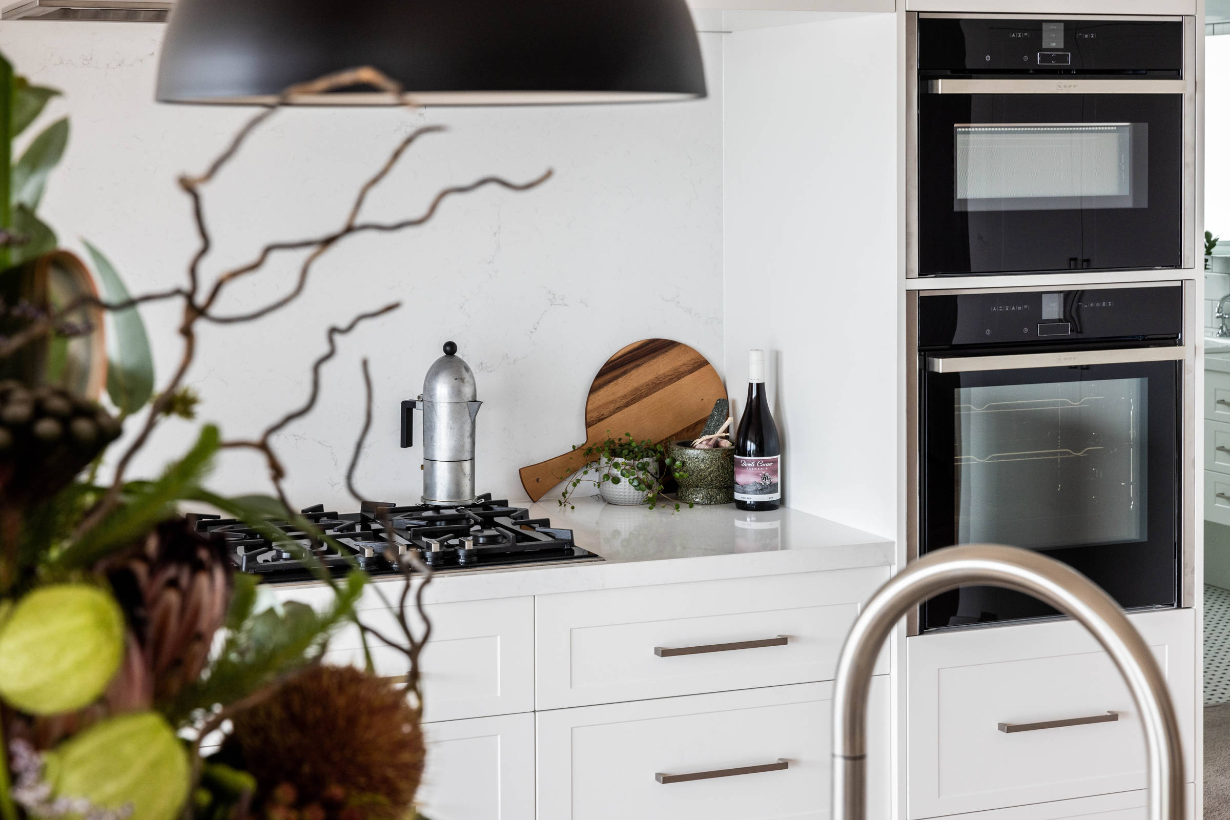 An Italian coffee pot, wooden chopping board, Tasmanian wine, garlic, and a mortar and pestle are right at home in this light and bright, contemporary kitchen renovation featuring solid stone benchtops, pendant lighting and brushed chrome tapware. Credit: Jordan Davis.