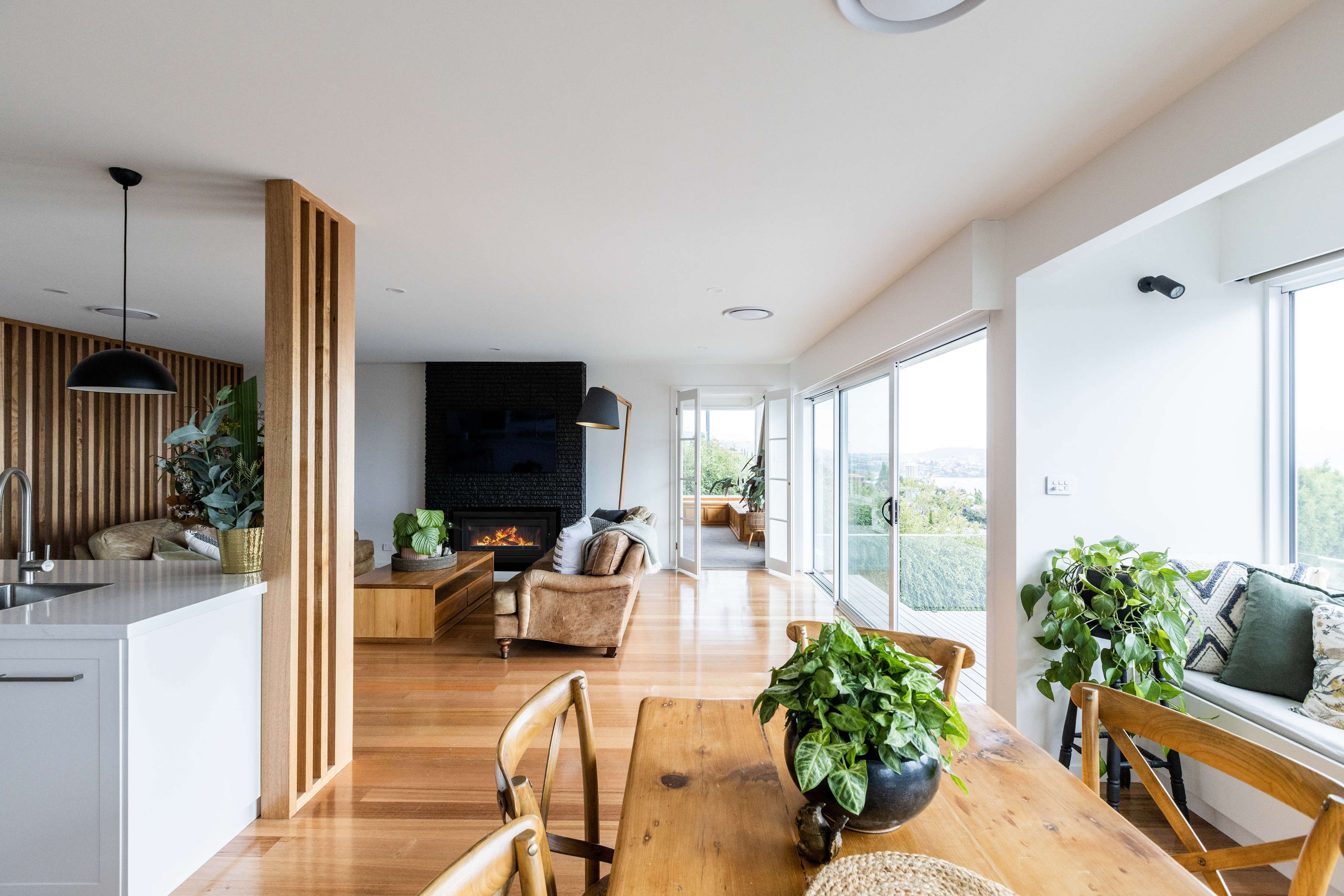 Kitchen and living room renovation, featuring Tasmanian oak flooring, solid stone benchtops, pendant lighting and other contemporary lighting. Our teams built to joinery standard Tasmanian oak screen and tasmanian oak feature joinery. The exising fireplace was painted to make it a feature and highlight one of the existing features of this 1970s house renovation. The result is very contemporary and light and bright and flows onto the deck extension at the front of the house. Credit: Jordan Davis.