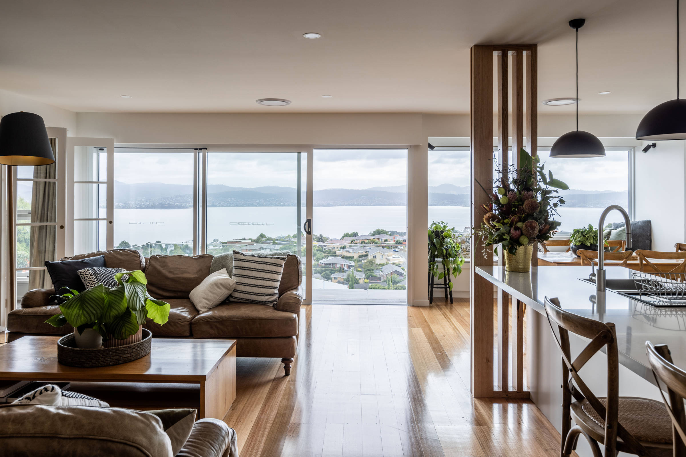 Kitchen and living room renovation, featuring Tasmanian oak flooring, solid stone benchtops, pendant lighting and other contemporary lighting. Our teams built to joinery standard Tasmanian oak screen and Tasmanian oak feature joinery. The result is very contemporary and light and bright and flows onto the deck extension at the front of the house. Credit: Jordan Davis.