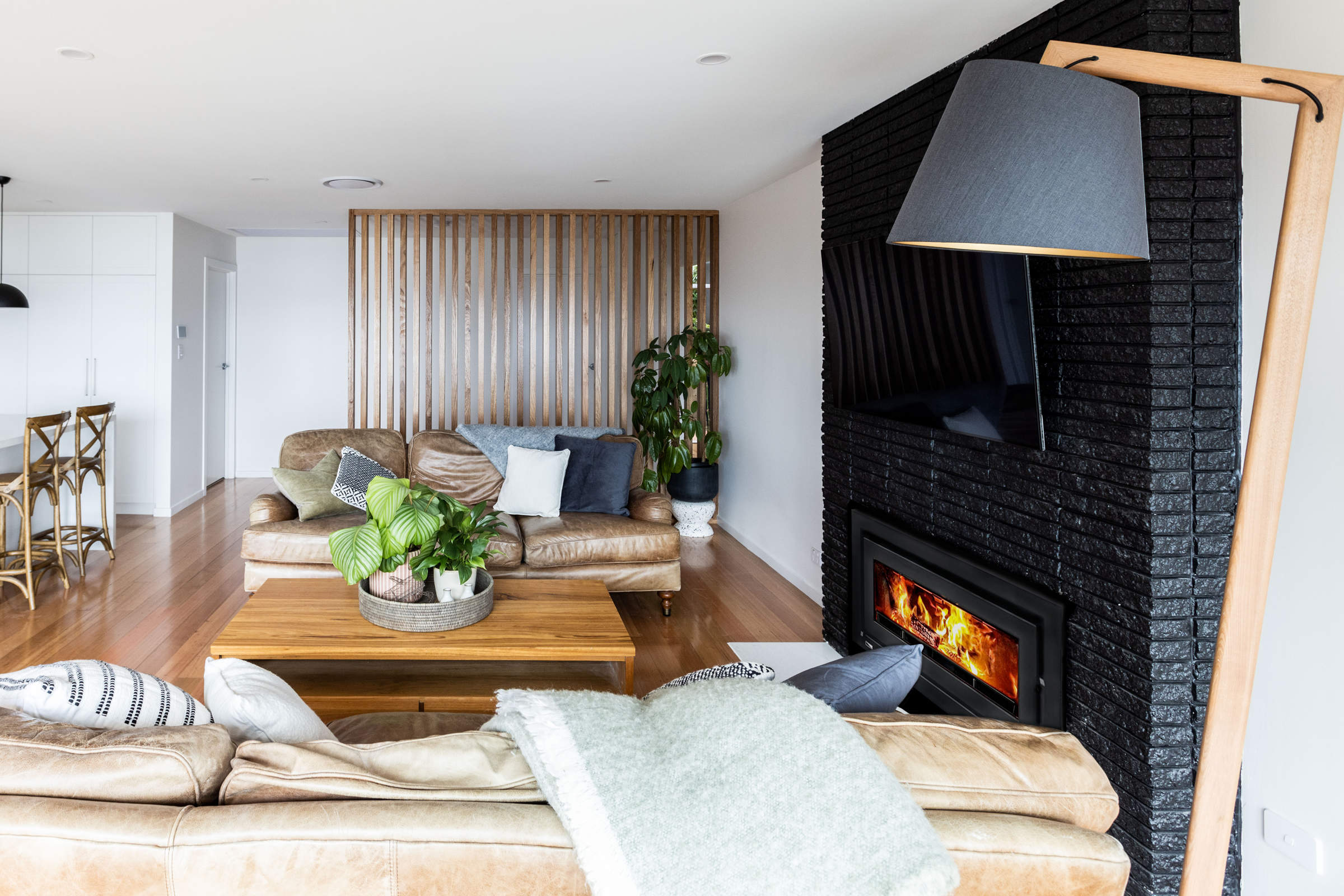 Kitchen and living room renovation, featuring Tasmanian oak flooring, solid stone benchtops, pendant lighting and other contemporary lighting. Our teams built to joinery standard Tasmanian oak screen and Tasmanian oak feature joinery. The exising fireplace was painted to make it a feature and highlight one of the existing features of this 1970s house renovation. Credit: Jordan Davis.