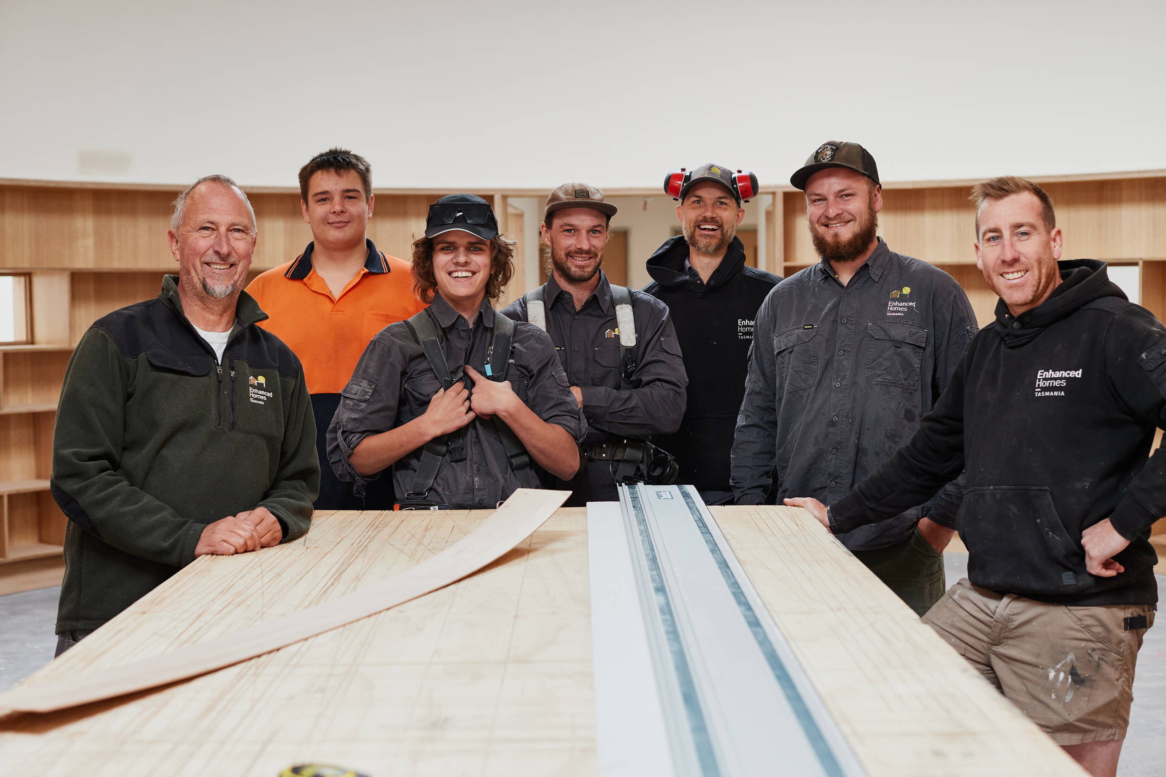 Seven male builders who are team members of the Enhanced Construction work team are in uniform and gathered around a table, looking at the camera in a portrait style image. Credit: Samuel Shelley.