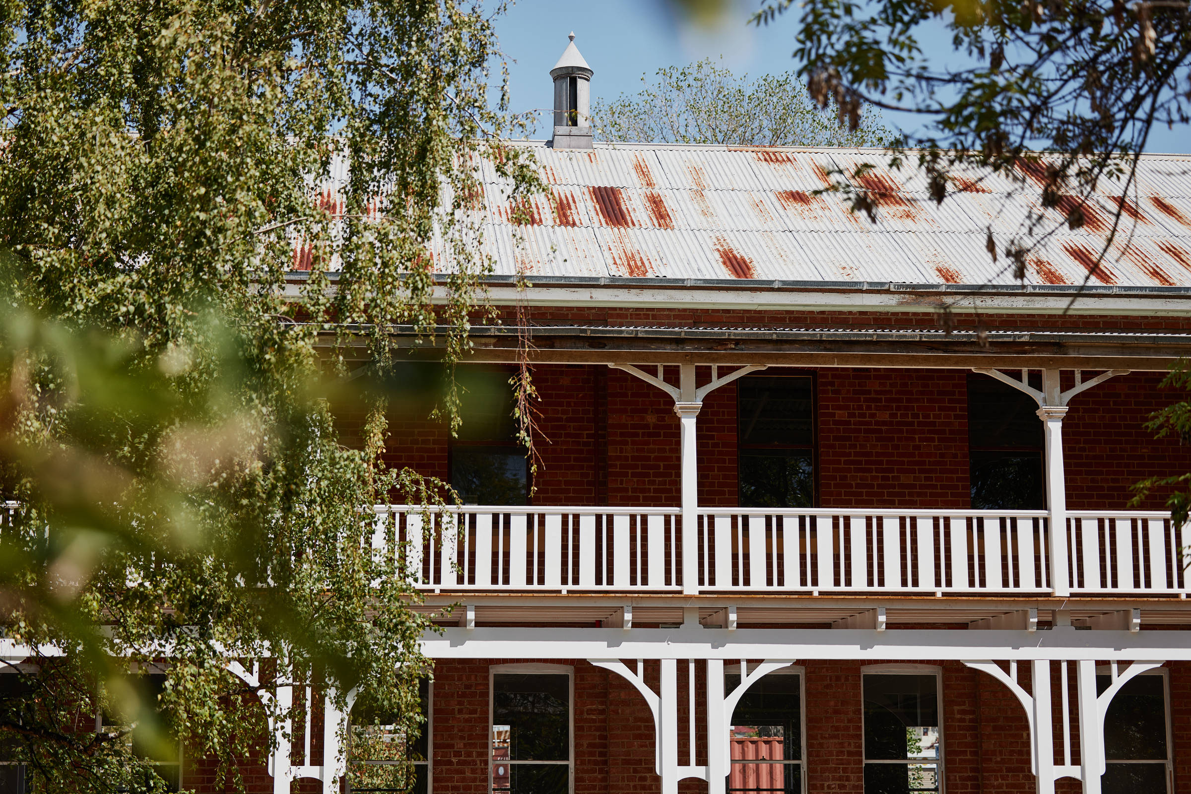 Partial image of a double storey heritage building with a slightly rusted roof and a freshly painted white verandah with decorative supports and balustrades, partially obscured by a tree on the left of the image. Credit: Samuel Shelley.