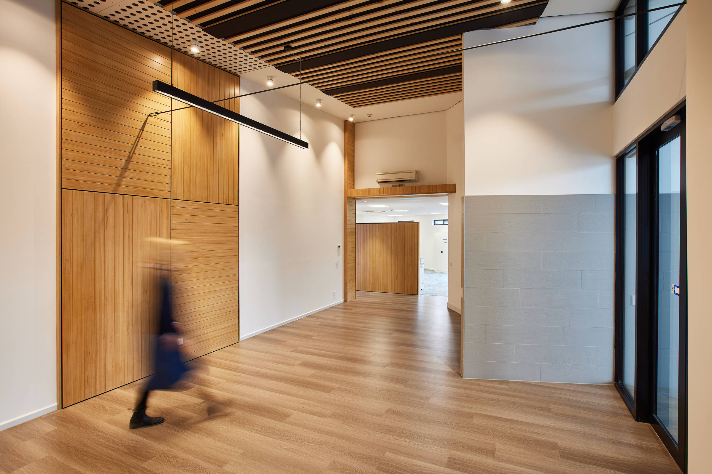 Blurred image of a woman walking through an entrance foyer. Tasmanian oak floor and detailing on the wall with black aluminium entrance doors, feature lighting and black and timber detailing on the ceiling. Credit: Samuel Shelley.