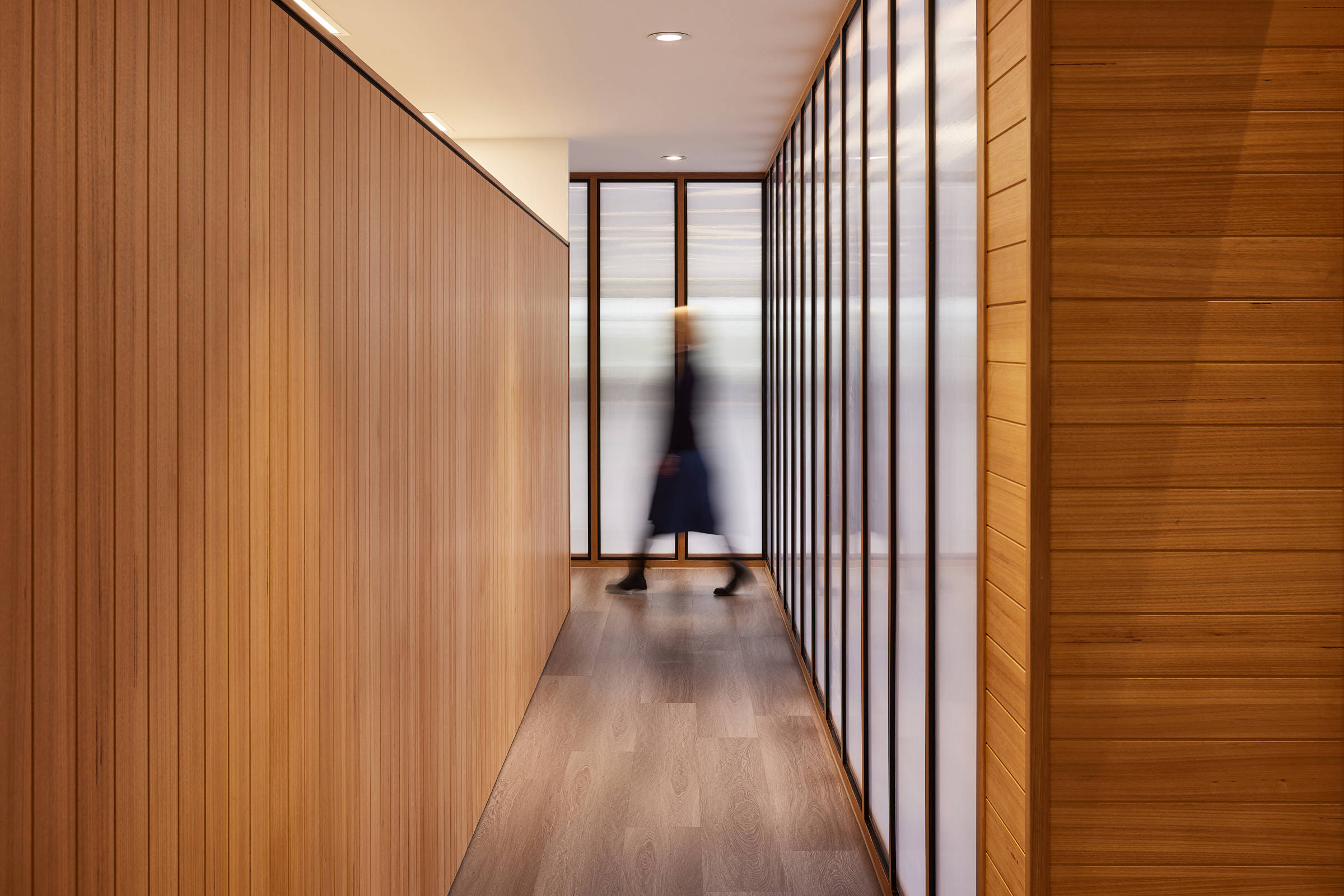 Blurred image of a woman walking through a hallway. Tasmanian oak floor and detailing on the wall and an internal decorative light wall of symmetrical panels, surrounded by black aluminium trim and Tasmanian oak on another. Credit: Samuel Shelley.