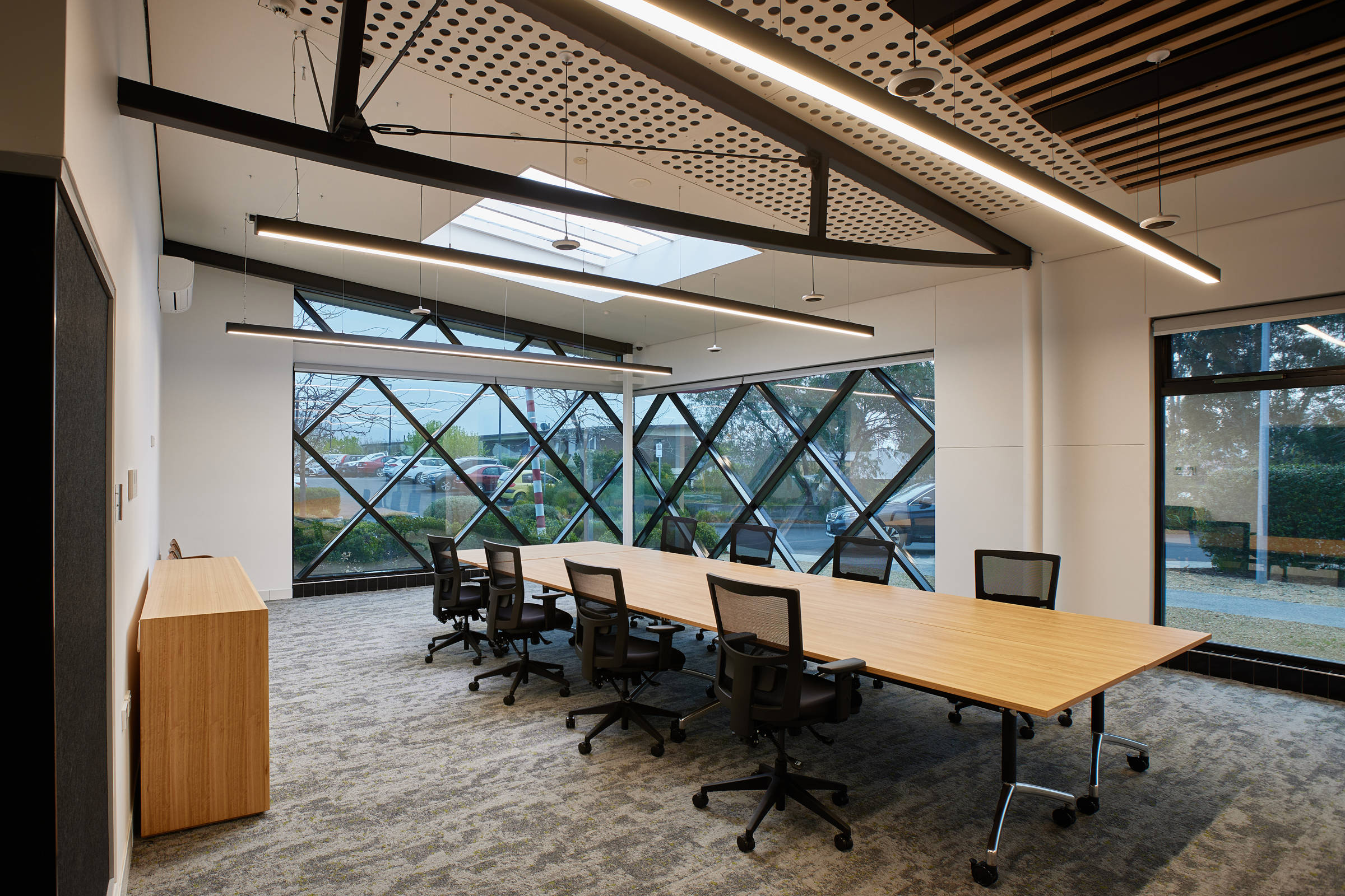 Large boardroom with a table, large black boardroom chairs, black architectural strip lighting hanging above the table and special triangular entire wall window detailing on two sides of the room. Credit: Samuel Shelley.