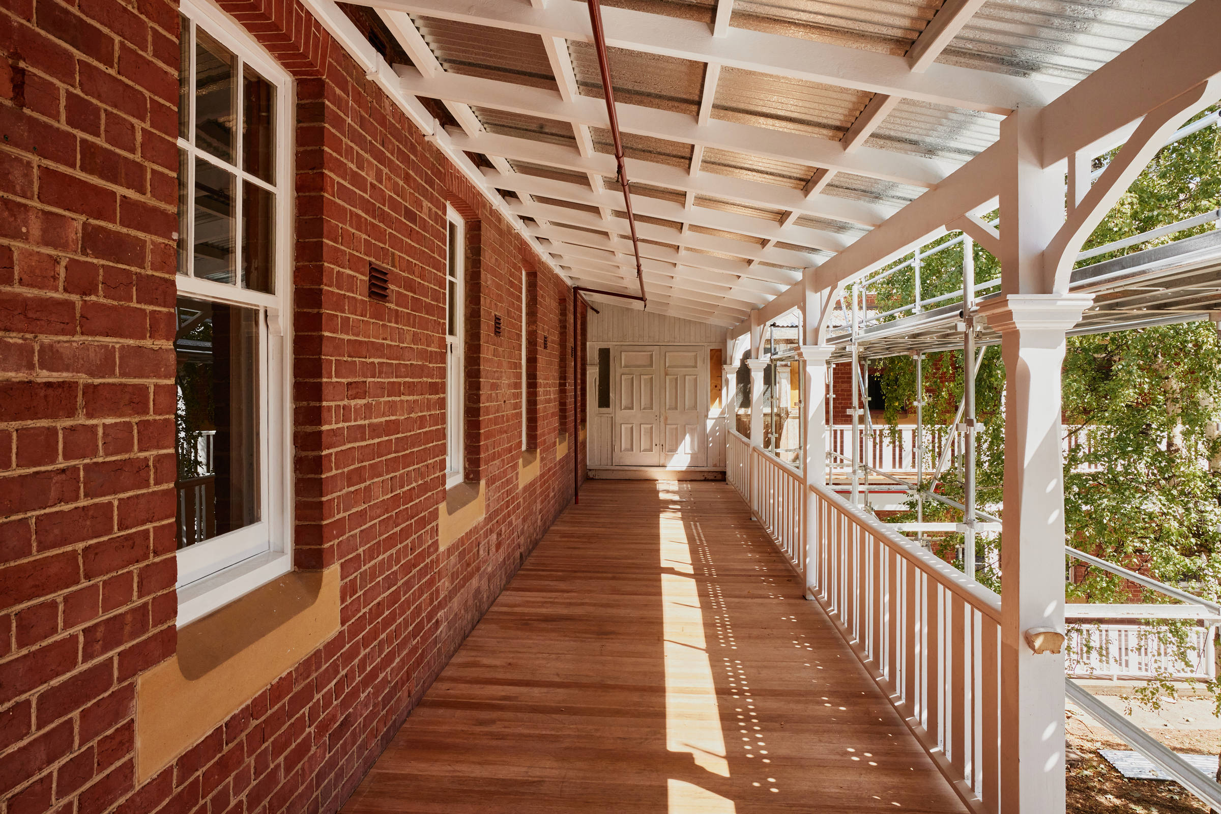 An 1860s redbrick heritage building second storey verandah. On the left side there are five white painted sash windows with sandstone sills. On the right side of the image is freshly painted decorative heritage balustrade, with shadows falling onto the new timber verandah floor through the still-erected scaffolding. At the end of the verandah are unrenovated double white doors. Credit: Samuel Shelley.