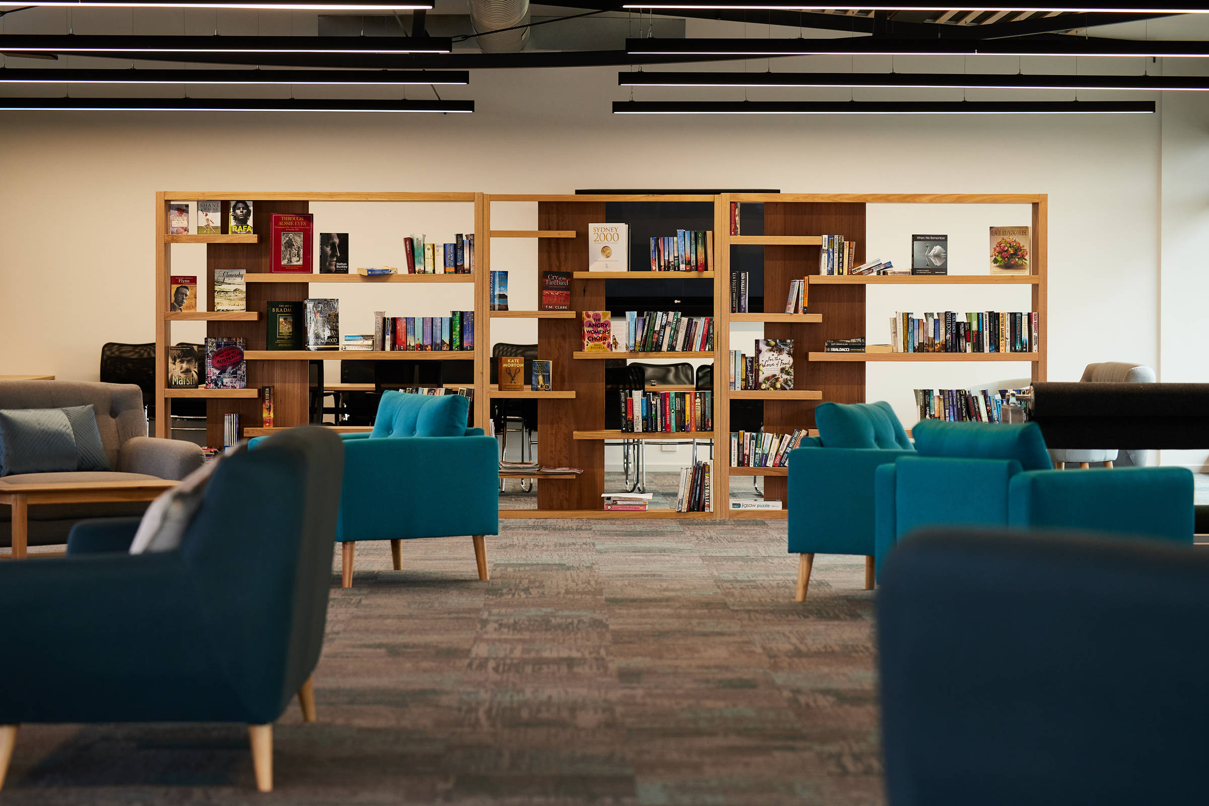 Internal view of a large timber bookshelf with offset shelves, stacked with books, surrounded by teal armchairs and prominent black architectural ducting on the ceiling. Credit: Samuel Shelley.