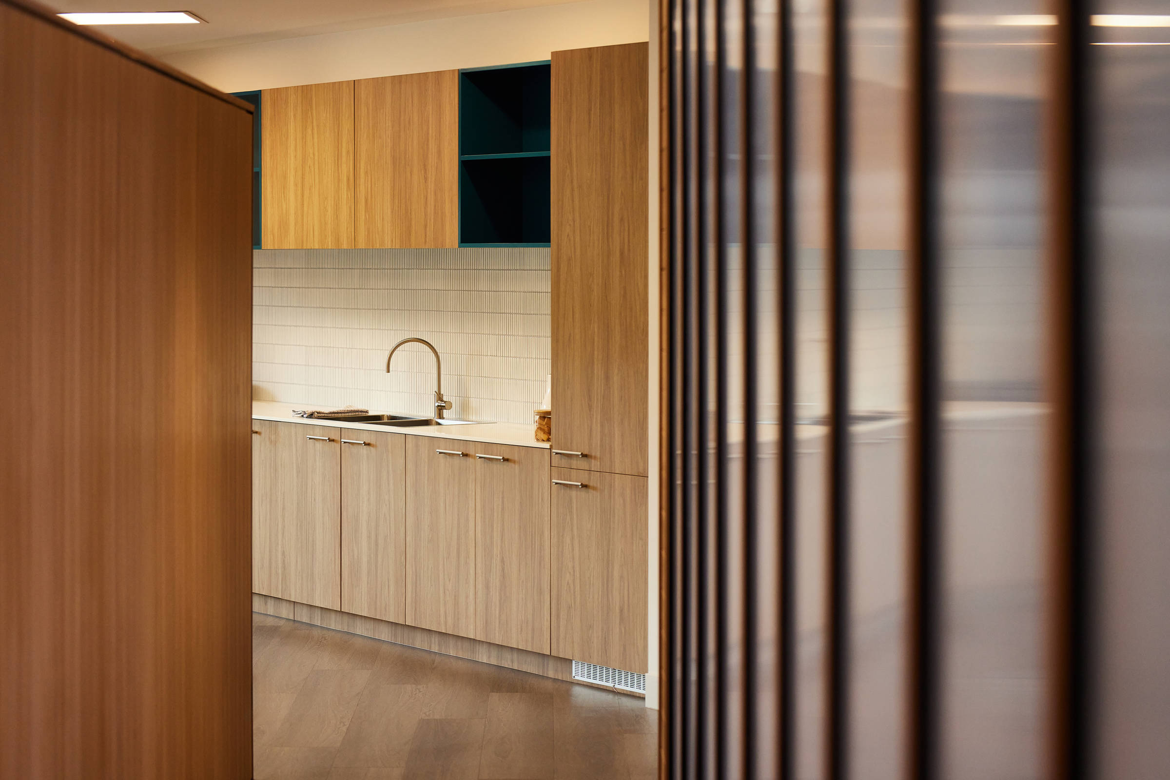 View of a small internal kitchenette with overhead and under sink cupboards and small vertically laid white tiles for the splashback. The right side of the image features the internal lightwall with black aluminium and timber detailing. Credit: Samuel Shelley.