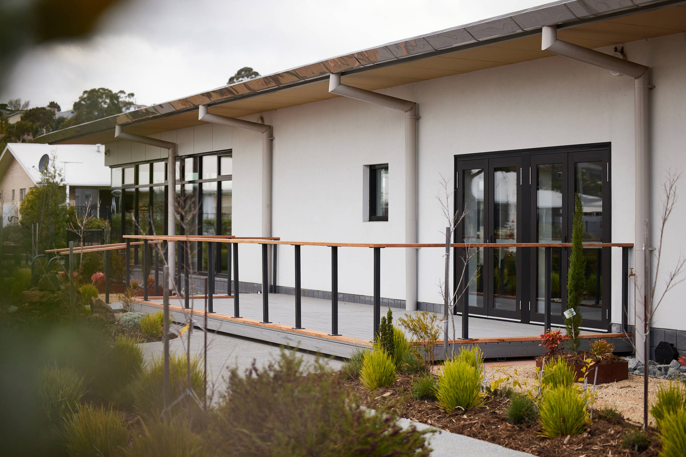 External view of black bi-fold doors leading to a grey tiled, ground level deck, with upright black posts and timber handrails, surrounded by a low level native garden. Credit: Samuel Shelley.