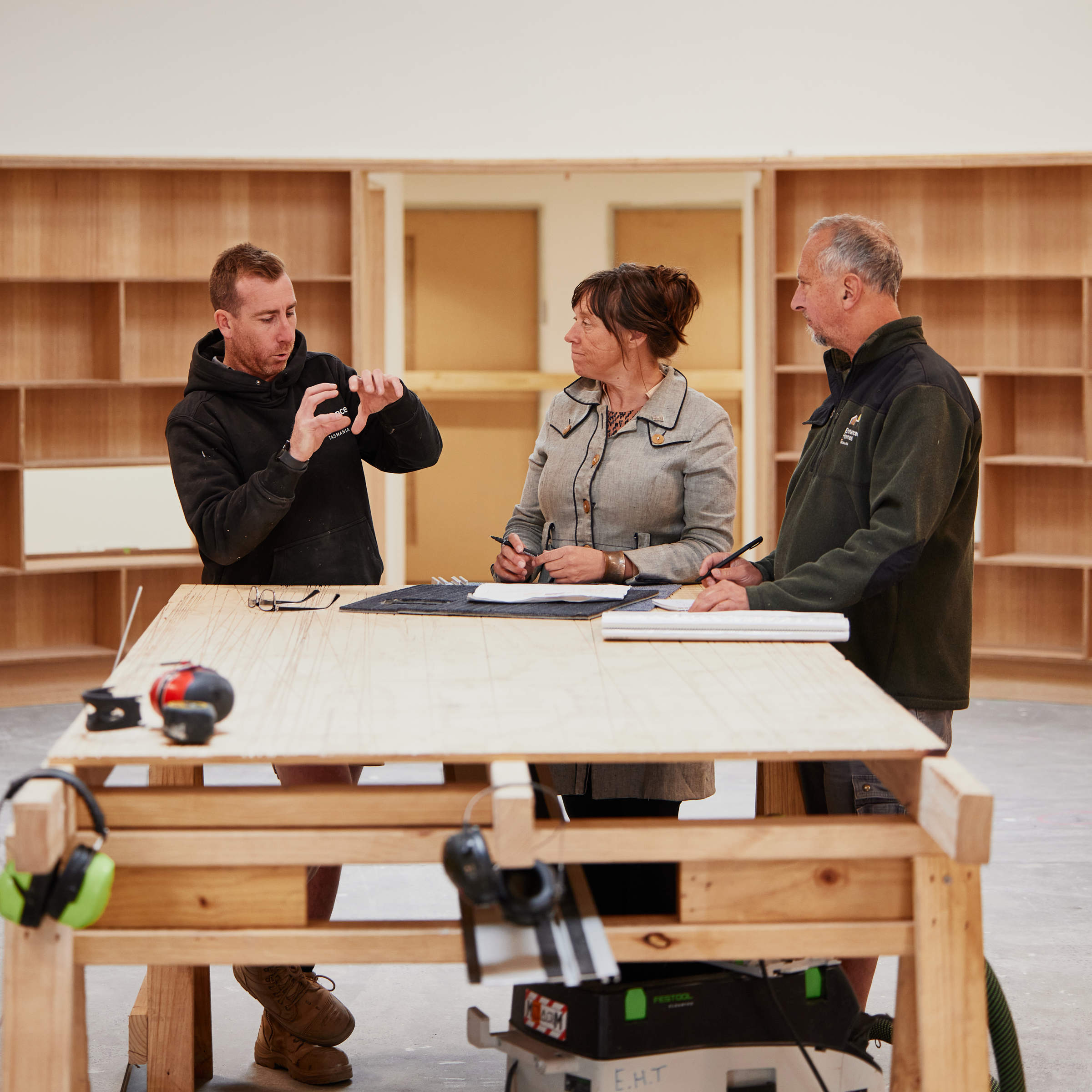 Male foreman, female architect and male project manager standing around a table and looking at building plans. The foreman is gesturing with his hands to discuss construction detail and the architect and project manager are looking at him. Credit: Samuel Shelley.