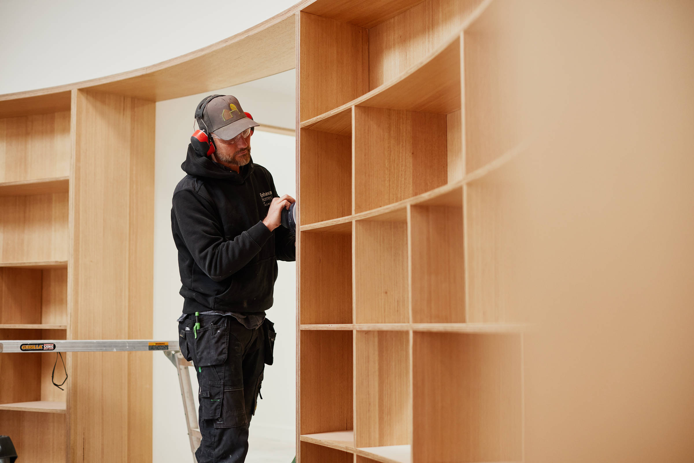 A male carpenter in his late 30s, doing detailed sanding work on the side of curved plywood bookshelves. Credit: Samuel Shelley.