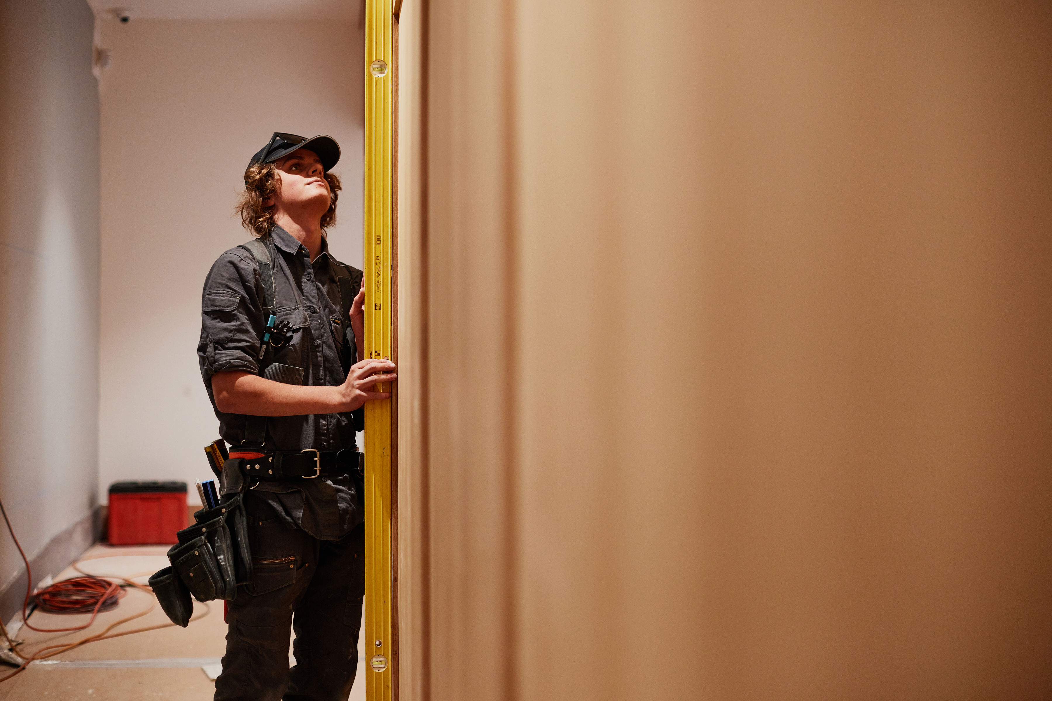 Young male apprentice builder holding a bright yellow spirit level vertically to measure the angle of a wall. Credit: Samuel Shelley.