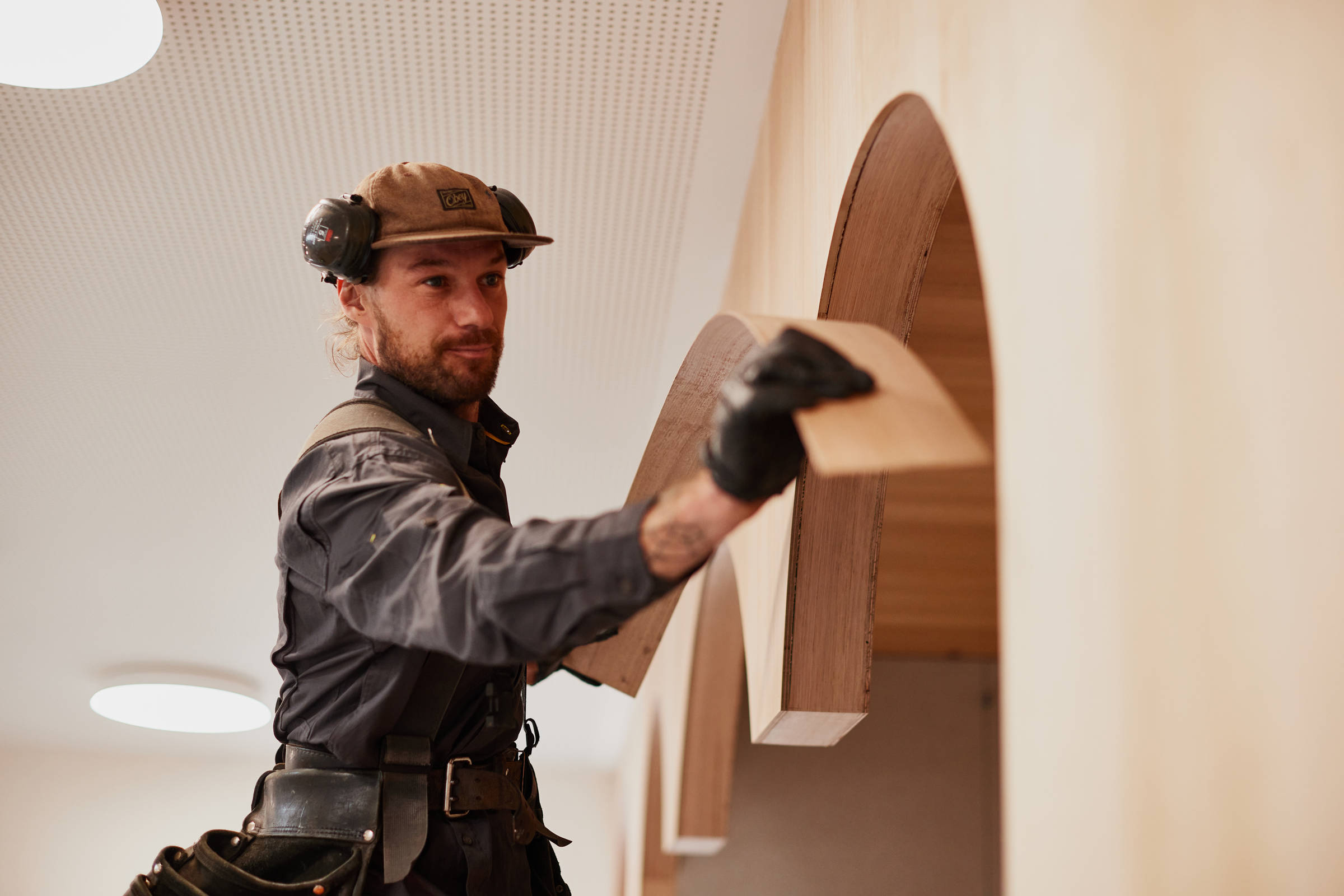 A male carpenter in his 30s who is wearing a cap, ear protection and a toolbelt is fitting curves of plywood into small timber arches into a book reading nook in a library. Credit: Samuel Shelley.