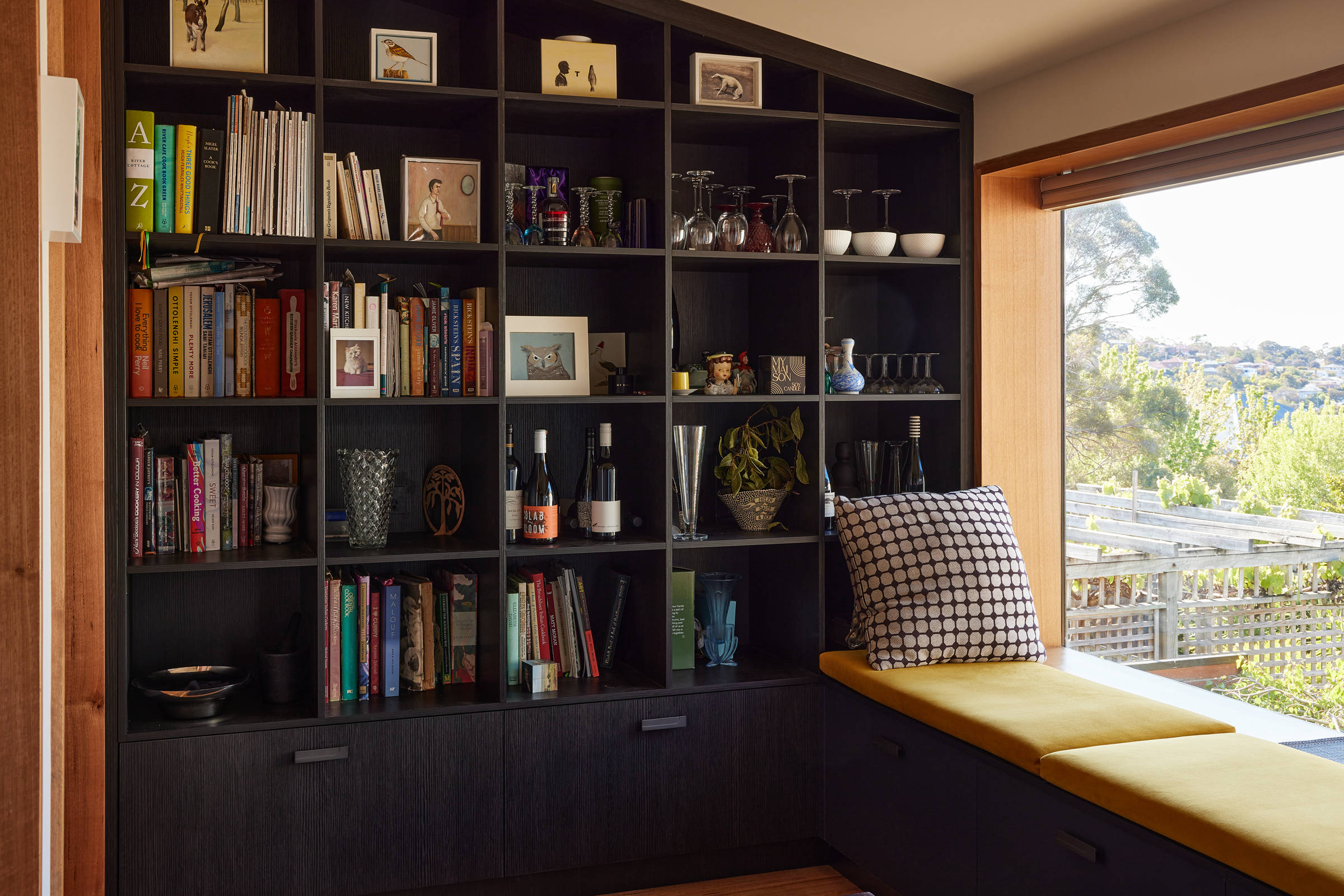 View onto a black pigeonhole style storage unit, with a windowseat and large window on the right of the picture. The pigeonholes are filled with artwork, books, glasses and ceramics. The windowseat cushions are mustard and there is a large black and white patterned cushion leaning against the wall. Credit: Samuel Shelley.