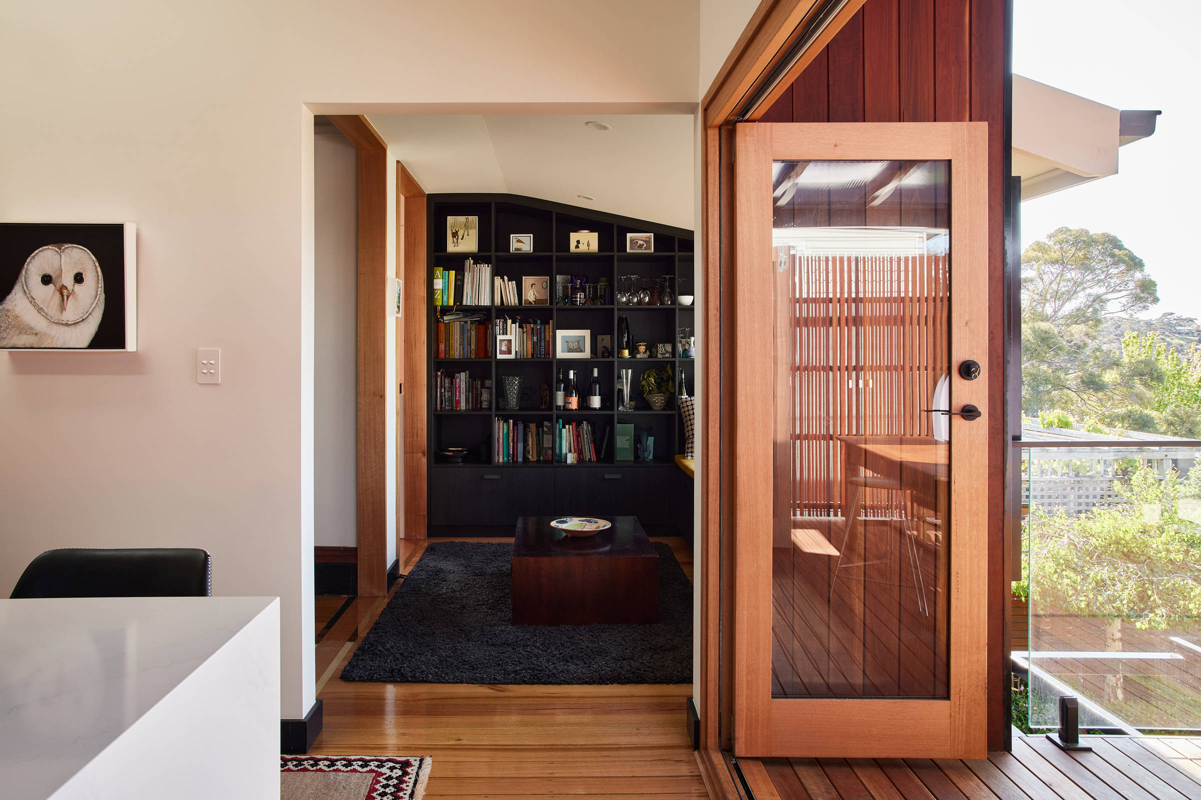 View onto a black pigeonhole style storage unit, looking over a dark coffee table and dark grey rug. The pigeonholes are filled with artwork, books, glasses and ceramics. The view is from the kitchen, looking through a wide open door frame with a timber door to the right. Credit: Samuel Shelley.