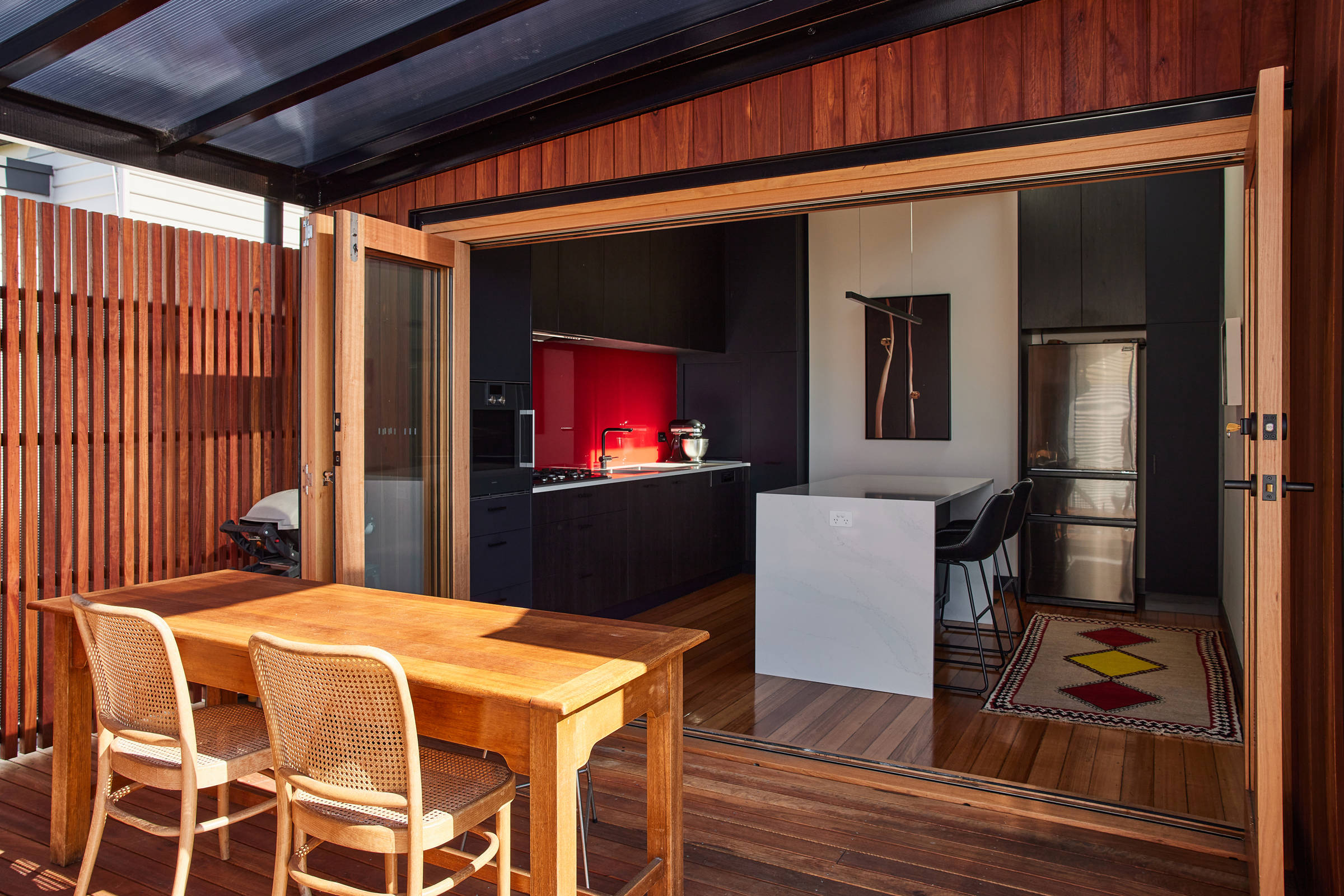 View from the deck looking back into the kitchen through bifold timber doors. There is a table and two chairs in the foreground and the kitchen features the main design elements of a bright red splashback, navy and black timber look cabinetry. There is a colourful rug in the kitchen, architectural lighting and one of the artist’s own paintings on the wall behind the island bench. Credit: Samuel Shelley.