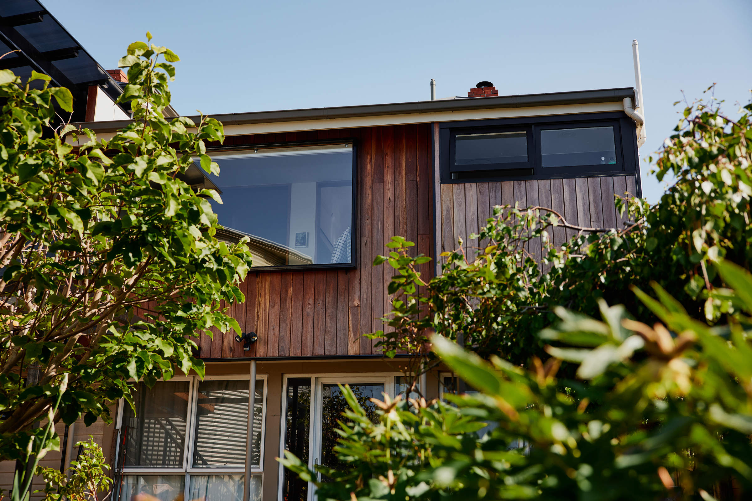 External view of a vertically timber clad house, looking back up onto the window seat and sitting nook. The windows are framed in black aluminium and trees from the garden frame the picture. Credit: Samuel Shelley.