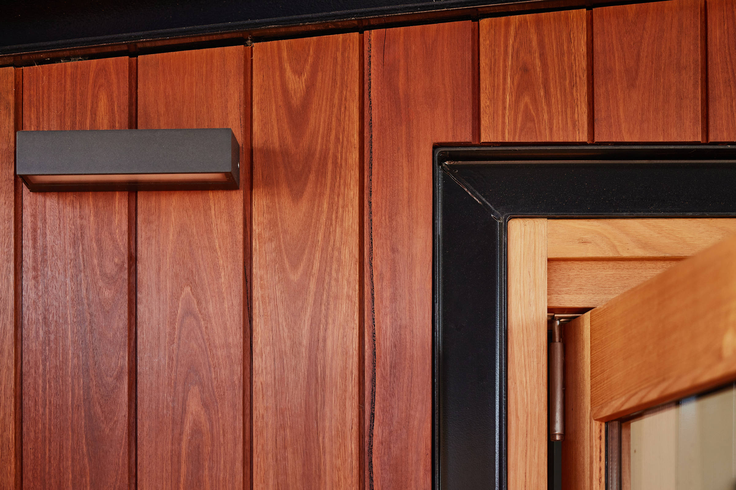 Closeup detail of the spotted gum external deck wall cladding with shiny black aluminium door surrounds and lighting. Credit: Samuel Shelley.