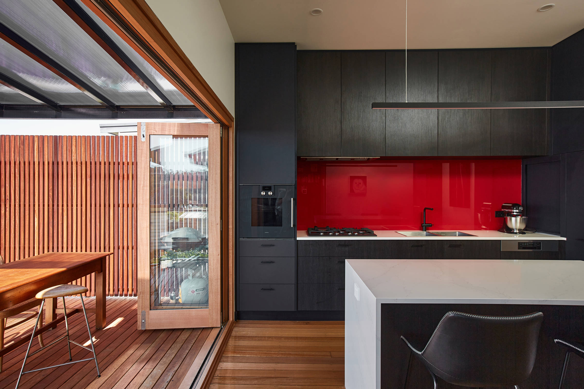 Kitchen leading onto a timber deck through timber framed bifold doors. The kitchen features black cupboards with a visible timber grain, soft grey benchtops and a bright red splashback. The single column floor to ceiling cabinetry to the right of the red splashback is in a dark navy blue. There is an island bench with the same soft grey benchtop. The deck features upright spotted gum narrow timber panels in a symmetrical pattern and a timber table in the same timber tones. Credit: Samuel Shelley.