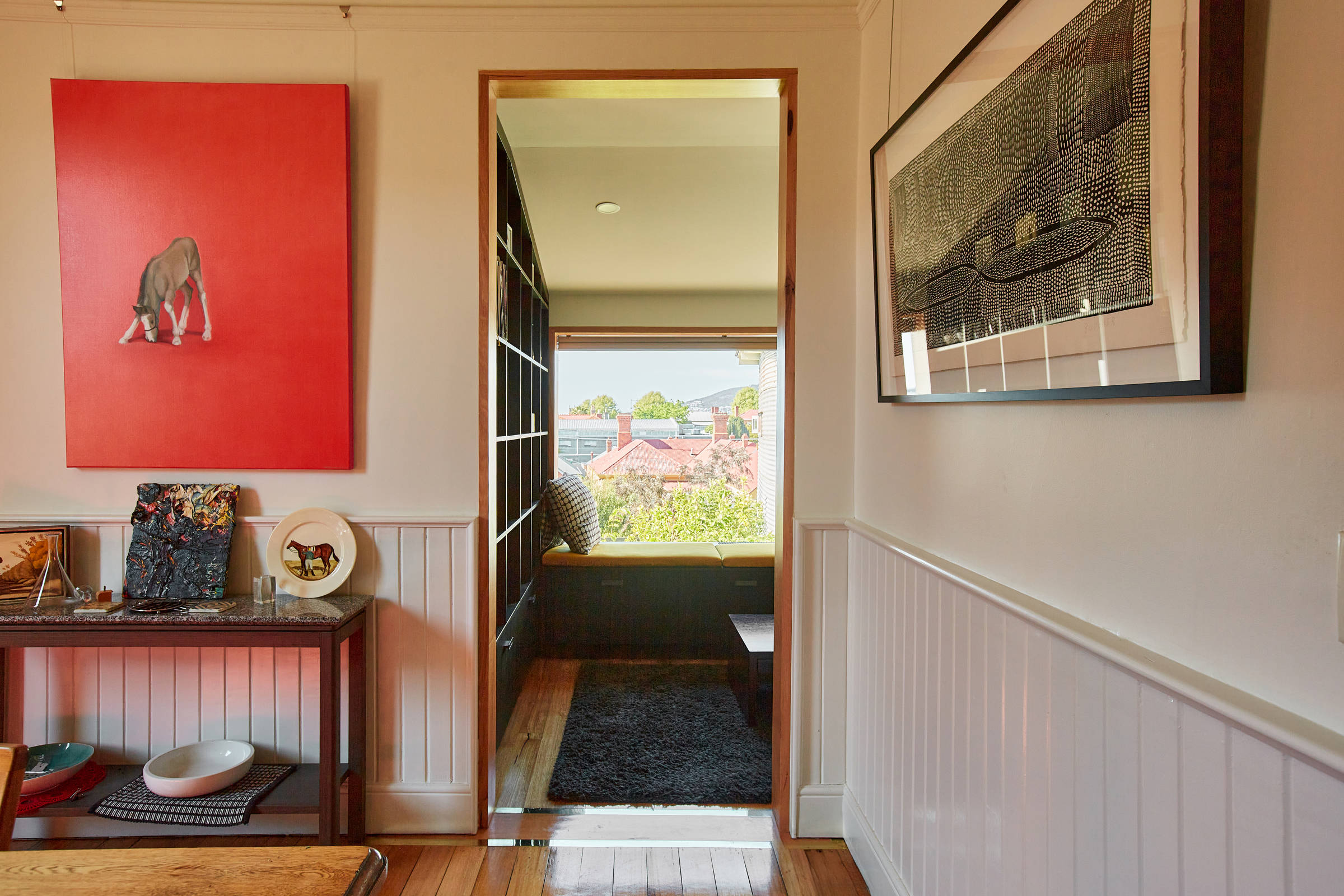 View looking south through a doorway over polished timber floors with a grey rug and a window seat with a mustard seat cover and a black and white cushion. To the left of the doorway is a small table with artwork and ceramics. The floor of the doorway has inlaid brass panels to make a highlight. Credit: Samuel Shelley.